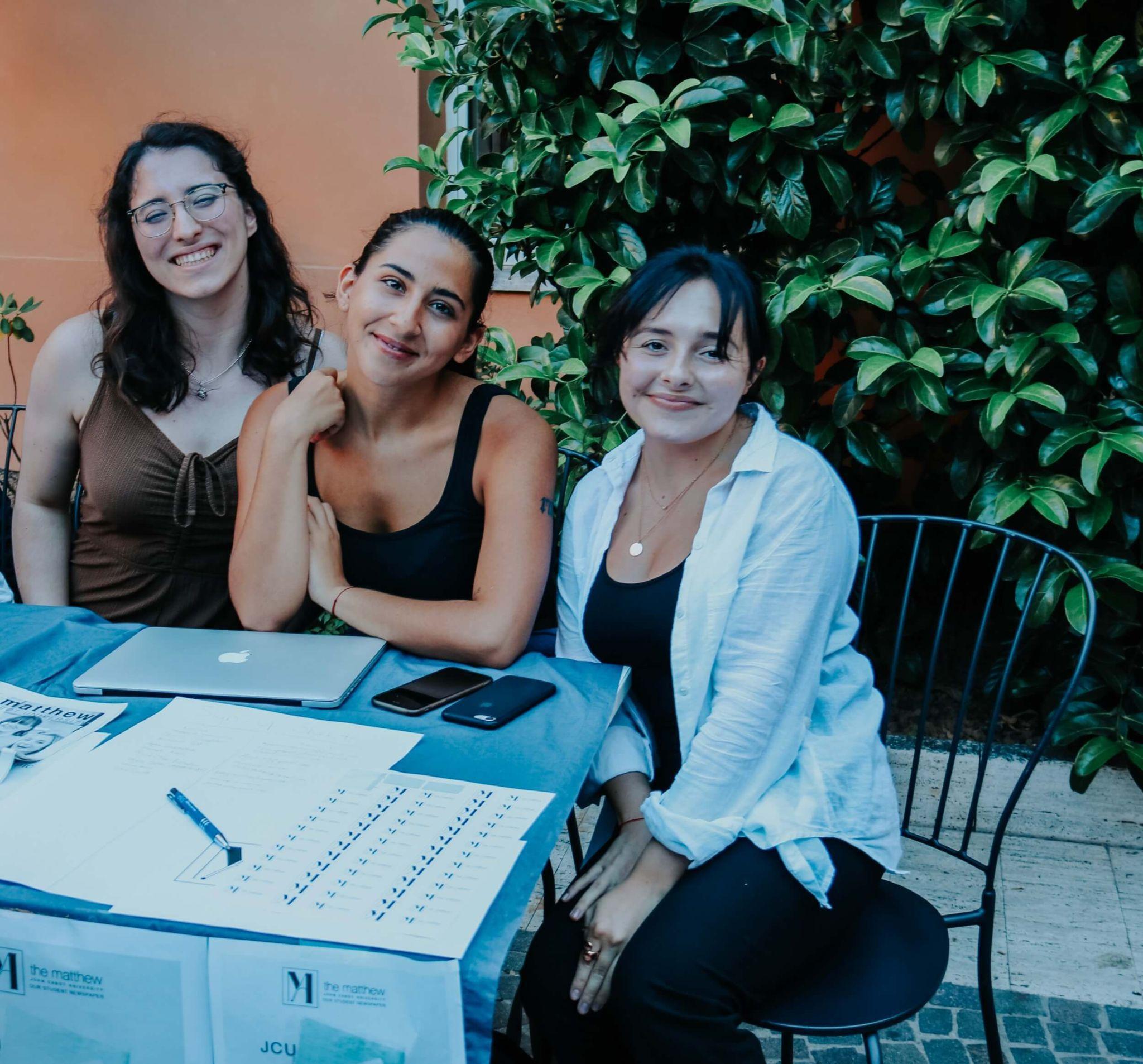 Three students smile at a registration table with materials, representing vibrant community life during their study abroad in Rome.