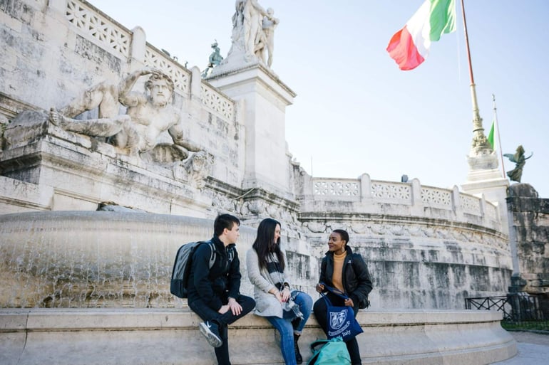 three students attending university in Rome sitting on a fountain and having a discussion.