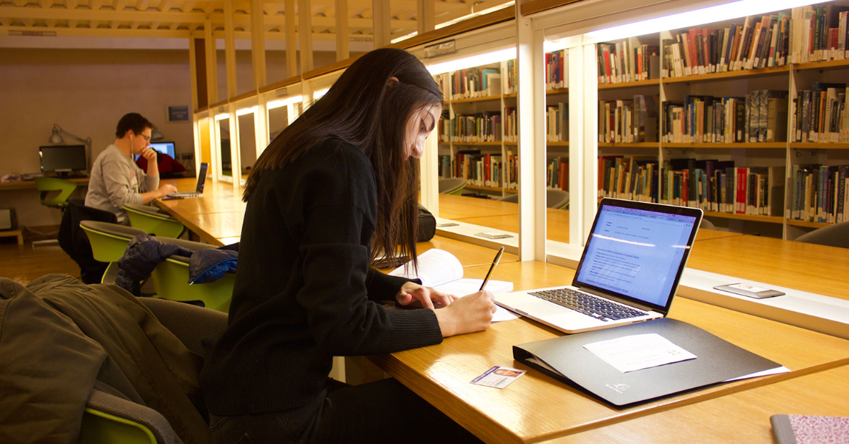 Girl sitting in library studying with a notebook and laptop.