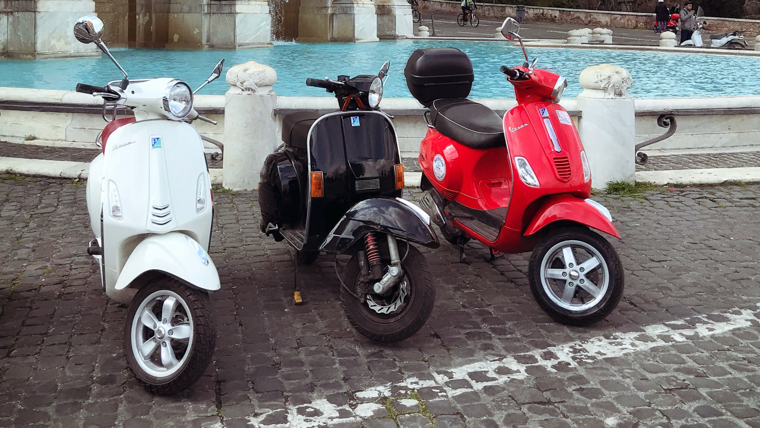 Three scooters lined up side by side in front of a fountain