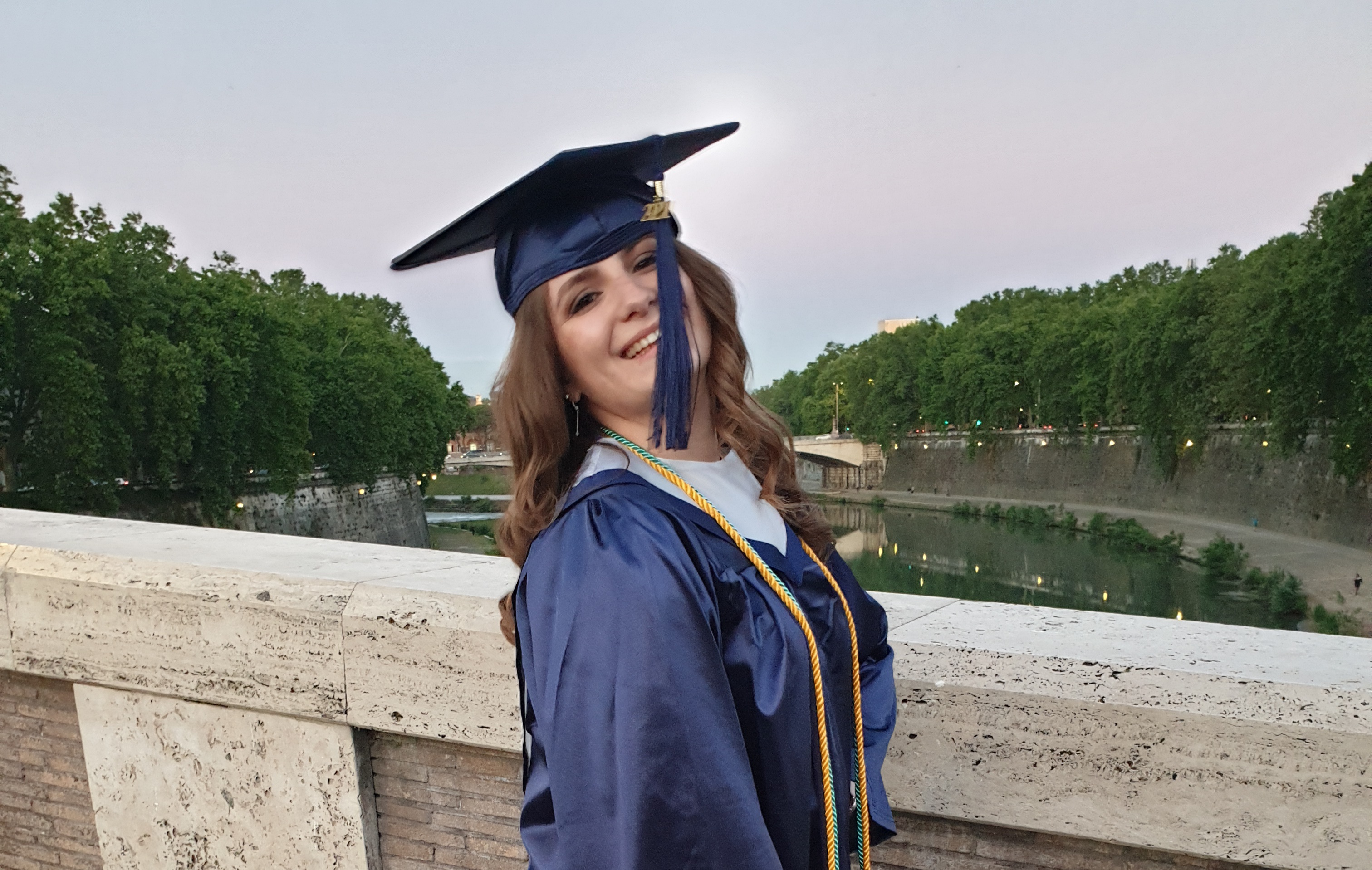 Ragazza riccia, con tunica blu appena laureata, sorride su un ponte a Roma 